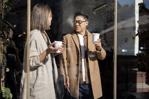 Asian businessman and caucasian businesswoman going, talking and drinking coffee inside unknown building. Concept of remote and freelance work. Idea of teamwork and business cooperation. Sunny day