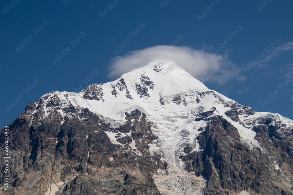 A panoramic view on the snow-capped peak of Tetnuldi in the Greater Caucasus Mountain Range in Georgia, Svaneti Region. A glacier is going down the mountain slopw. Sharp peaks, wanderlust, solitude.