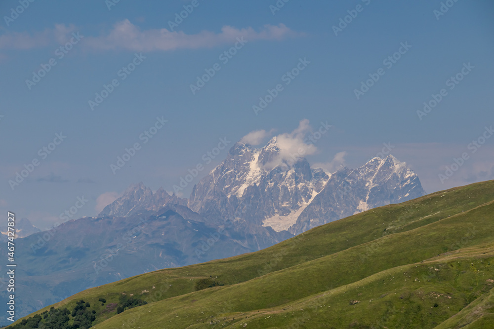 Lush green hills with panoramic view on high, sharp and snow-capped mountains in Greater Caucasus Mountain Range in Georgia, Upper Svaneti Region. Wilderness, remote location, solitude.