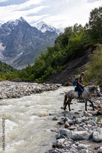 Woman crossing river Adishischala with a horse. View on Adishi Glacier and on the snow-capped peaks of Tetnuldi, Gistola and Lakutsia in the Greater Caucasus Mountain Range in Georgia, Svaneti Region.