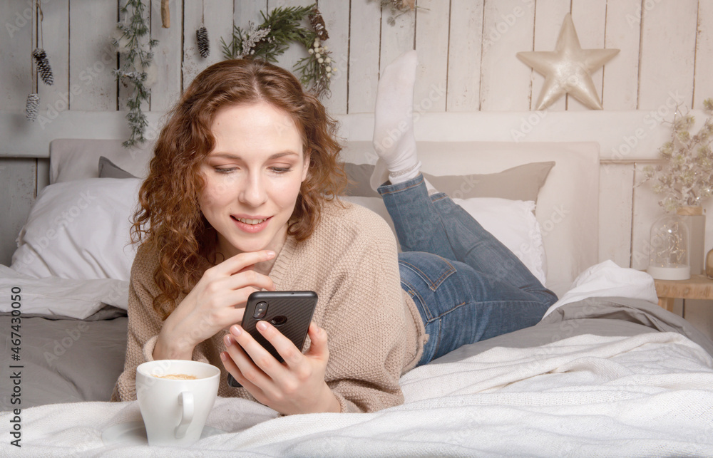 Beautiful caucasian girl lyes on the bed and looks in the phone with cup of coffee christmas Beautiful caucasian girl lyes on the bed and looks in the phone with cup of coffee christmas