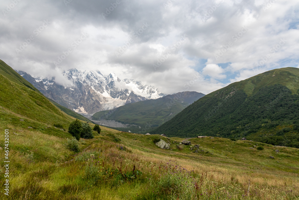 A panoramic view on the snow-capped peaks of Tetnuldi, Gistola and Lakutsia in the Greater Caucasus Mountain Range in Georgia, Svaneti Region. Hills with lush pastures, peaks covered in clouds.
