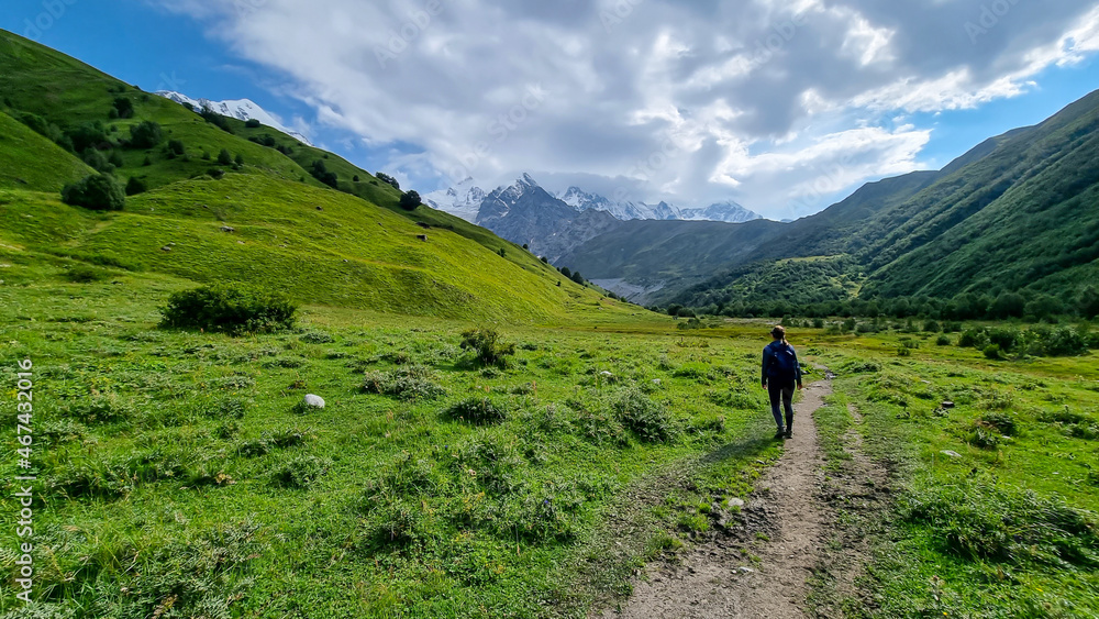 A woman hiking through a valley with a view on the snow-capped peaks of Tetnuldi, Gistola and Lakutsia in the Greater Caucasus Mountain Range in Georgia, Svaneti Region. Wanderlust. A woman hiking through a valley with a view on the snow-capped peaks of Tetnuldi, Gistola and Lakutsia in the Greater Caucasus Mountain Range in Georgia, Svaneti Region. Wanderlust.