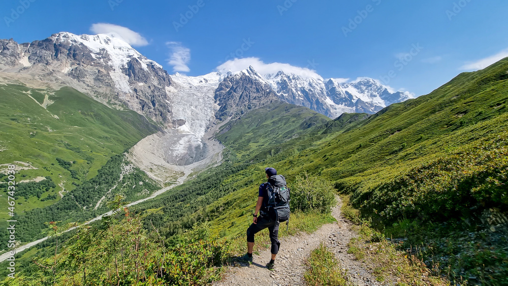 A man on a hiking trail with a panoramic view on the snow-capped peaks of Tetnuldi, Gistola, Lakutsia and the Adishi Glacier in the Greater Caucasus Mountain Range in Georgia,Svaneti Region.Wanderlust