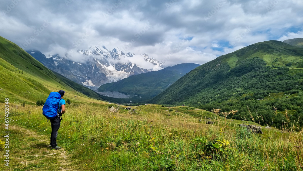 A man hiking  with a view on the snow-capped peaks of Tetnuldi, Gistola and Lakutsia in the Greater Caucasus Mountain Range in Georgia, Svaneti Region. The backpack is protected by a rain cover.