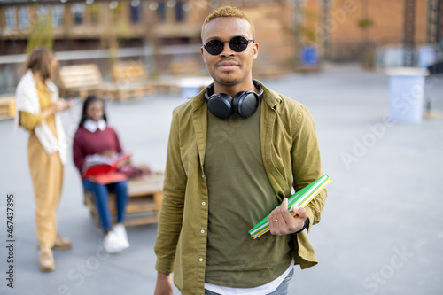 Smiling black student guy on background of her female friends outdoors. Concept of education and learning. Idea of student lifestyle. Young man holding books and looking at camera. University campus