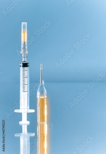 A syringe with a burning needle and divisions next to a glass ampoule with a gold glowing filling, vertically on a blue background.