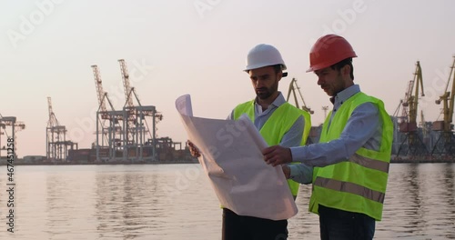Engineers wearing helmets and reflective vests regulate building of cargo cranes using delineation standing on beach against port