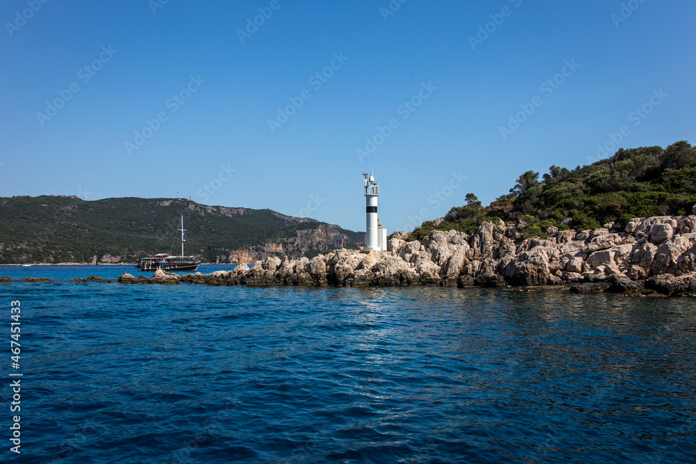 A beautiful small lighthouse on the edge of an island over the rocks and blue Mediterranean sea, at the coast of Turkey, a boat passing by behind with a big green mountain in the background.