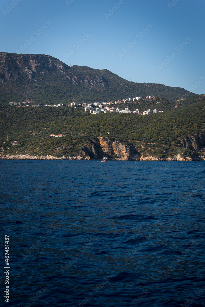 A beautiful green mountain and a city with white houses on top while a sailing boat moving through the rocky shore on the blue Mediterranean waters near by the coast of Kas, Antalya, Turkey. 