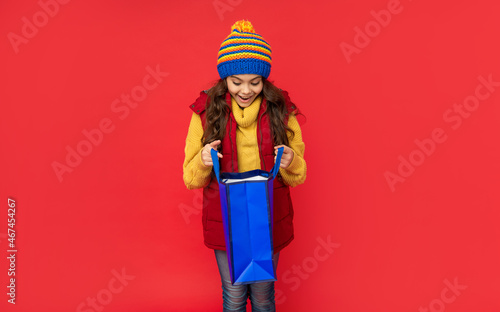 amazed child in winter clothes open shopping bag on red background, christmas shopping