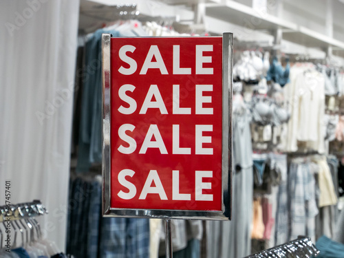 A red advertising sign in a store with the inscription sale, against the background of hangers with clothes. Sale in a retail clothing store, in a boutique, in a shopping center.