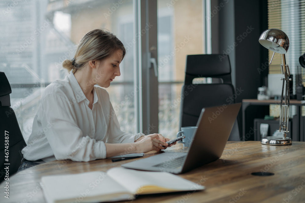 Young woman with laptop and phone at work table in modern office in the afternoon