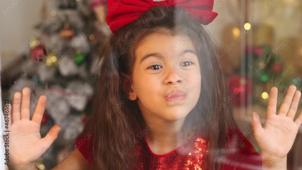 portrait of a little cute girl in a red dress with a bow on her head through glass close up. Merry Christmas and Happy Holidays. Happy New Year. Hygge. Winter. High quality 4k footage portrait of a little cute girl in a red dress with a bow on her head through glass close up. Merry Christmas and Happy Holidays. Happy New Year. Hygge. Winter. High quality 4k footage