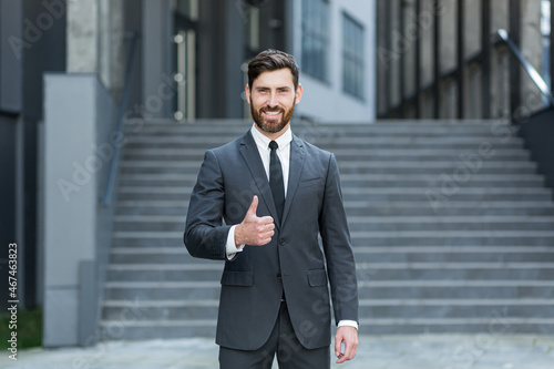 Portrait of young successful businessman looking at camera and smiling outdoors holding thumbs up rejoicing