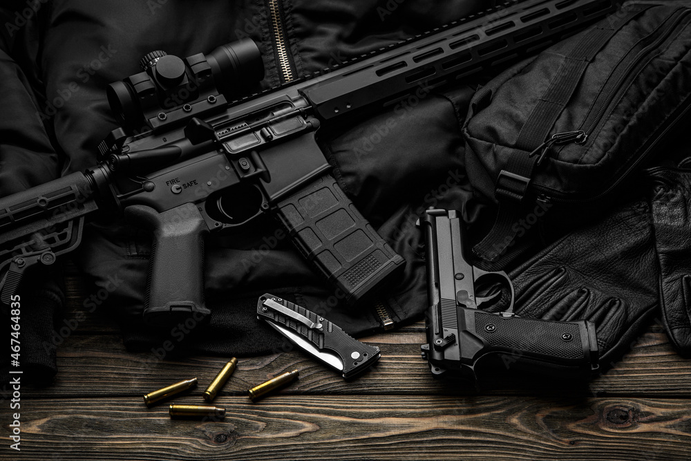 Modern automatic rifle with a telescopic sight on a dark background. Jacket, cap, gloves on a wooden table. The uniform of a guard or a mercenary Modern automatic rifle with a telescopic sight on a dark background. Jacket, cap, gloves on a wooden table. The uniform of a guard or a mercenary