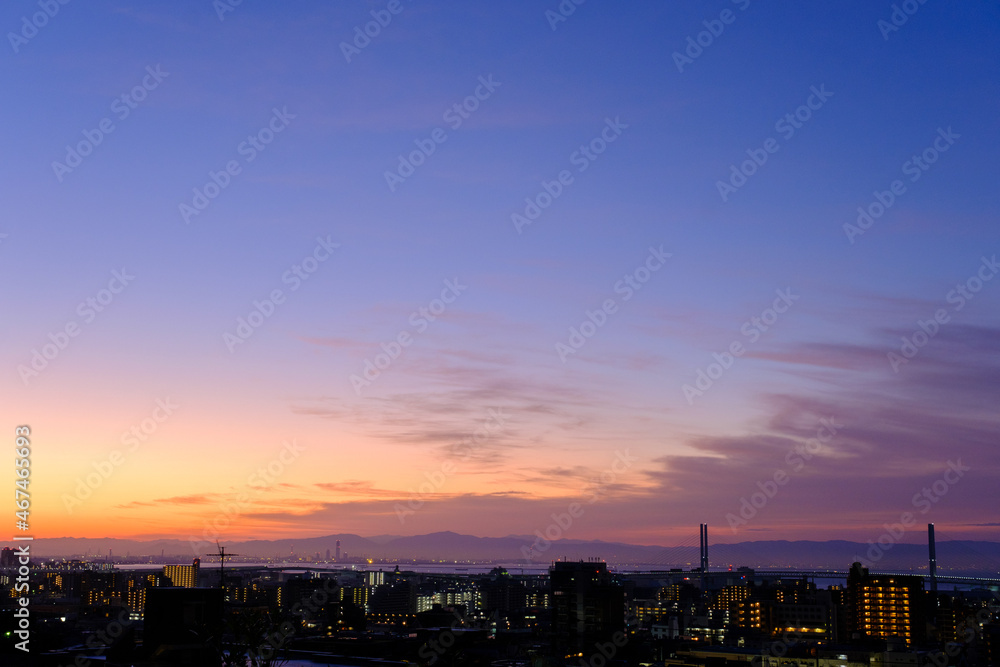 都市の夜明け前。日の出とともに空と雲がオレンジ色に染まる。遠くに大阪湾を臨む