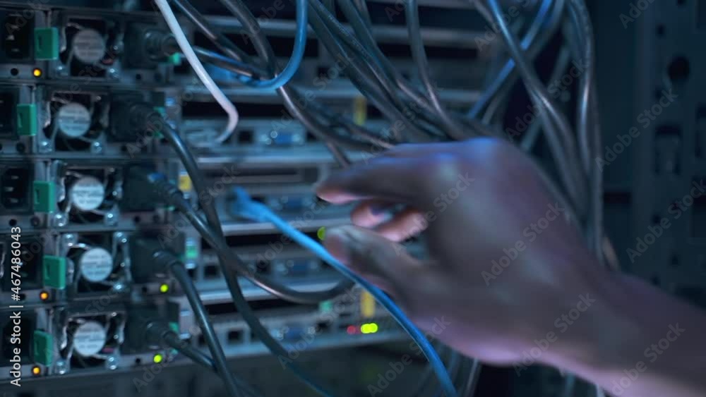 Engineer specialist works in server room. African-American employee switches cables fixing network equipment spbas on rack in semi-dark data center closeup
