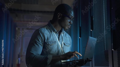 Engineer serving of contemporary data center. African-American person with glasses spbas illuminated by laptop screen sets up powerful servers at racks closeup