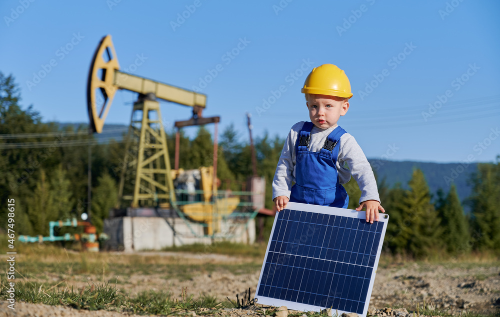 Portrait of young child guy standing on blurred background of oil rocking pump and holding in his hands small solar panel. Future power engineer demonstrating ecological way of obtaining energy.