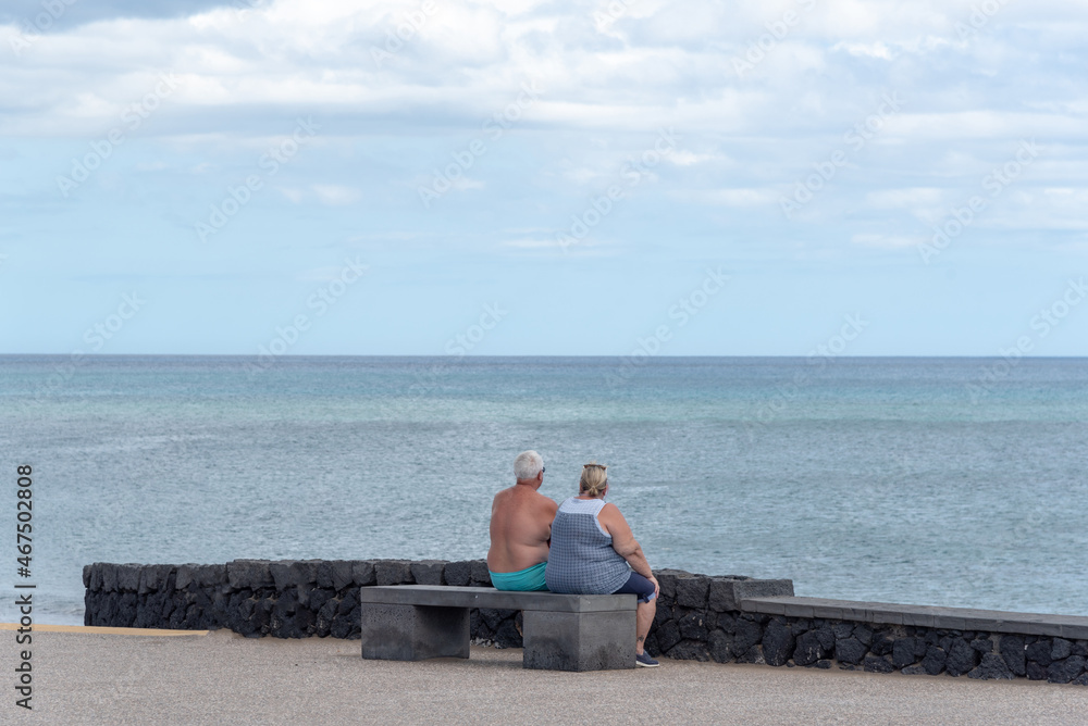 un pareja de personas mayores de espaldas sentado en un banco mirando el mar cerca de la playa. 