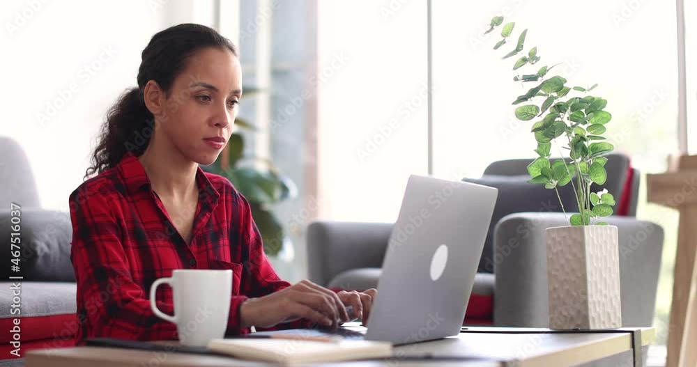 Ethnic female freelancer using laptop and making notes while sitting at table near sofa and working on remote project at home Ethnic female freelancer using laptop and making notes while sitting at table near sofa and working on remote project at home