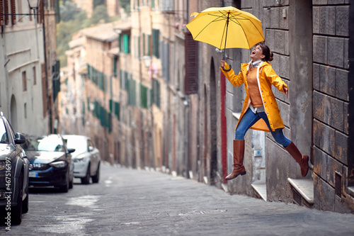 A young girl with a yellow raincoat and umbrella is jumping on the street while walking the city on a rainy day. Walk, rain, city
