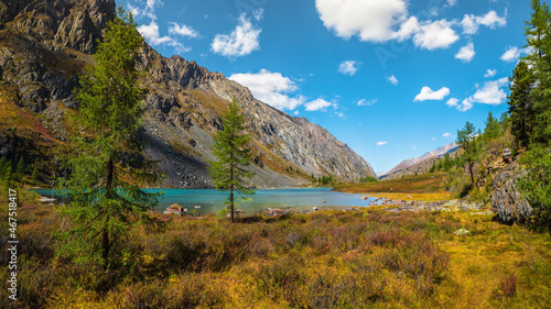 Fototapeta Naklejka Na Ścianę i Meble -  Panoramic alpine landscape with mountain lake in green valley in summer under blue sky. Awesome highland scenery with beautiful glacial lake among sunlit hills and rocks against mountain range.