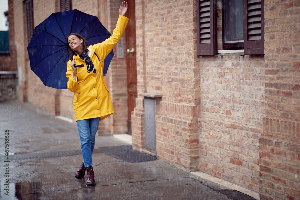 A young handsome woman in a yellow raincoat and with umbrella is dancing while walking the city on a rainy day. Walk, rain, city