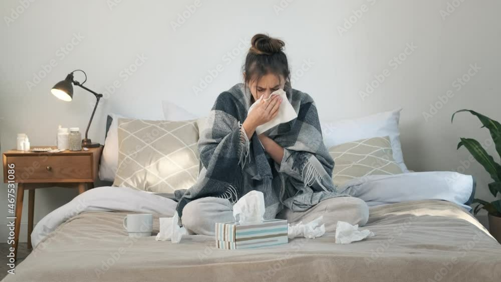 Sick young woman with fever blows her nose in a napkin, covered in blanket, having flu symptoms. Unhealthy girl in bed at home Sick young woman with fever blows her nose in a napkin, covered in blanket, having flu symptoms. Unhealthy girl in bed at home
