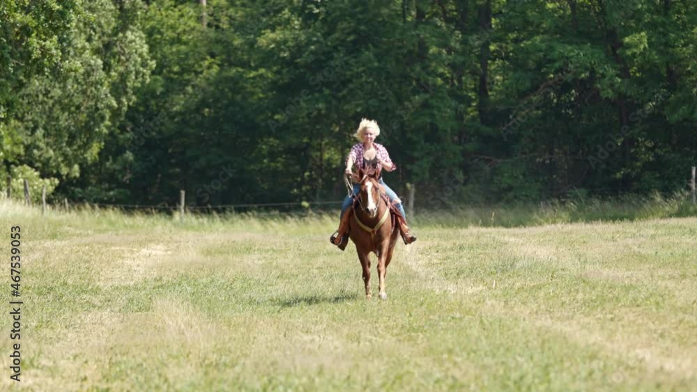 A cowgirl with blonde curls and a checked shirt rides a brown horse at a gallop towards the camera. A cowgirl with blonde curls and a checked shirt rides a brown horse at a gallop towards the camera.