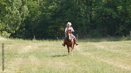 A cowgirl with blonde curls and a checked shirt rides a brown horse at a gallop towards the camera.  photo