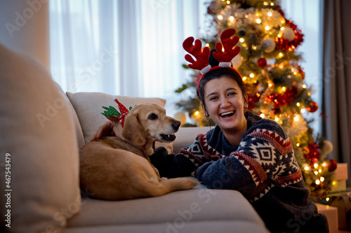 Happy young woman celebrating festive season with little yellow dog by christmas tree. Smiling girl and her dog wearing christmas antlers sitting on sofa at home