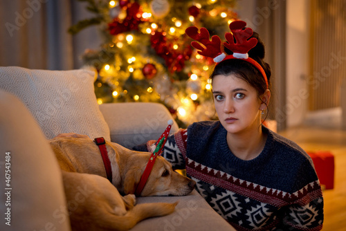 Sad lonely young woman sitting frustrated next to her yellow dog by couch at home during festive season, wearing Christmas antlers