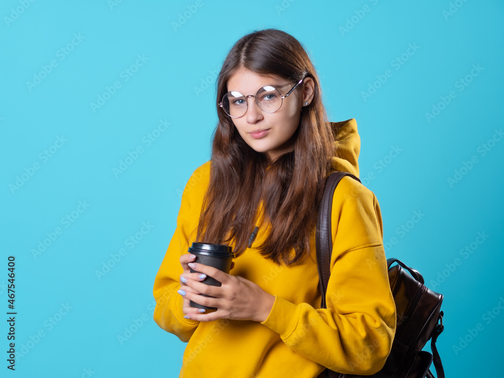 Cute young brunette in jeans and yellow hoodie. A beautiful student in glasses with a backpack on her shoulders holds a takeaway coffee in her hands, a studio portrait on a blue background. Cute young brunette in jeans and yellow hoodie. A beautiful student in glasses with a backpack on her shoulders holds a takeaway coffee in her hands, a studio portrait on a blue background.