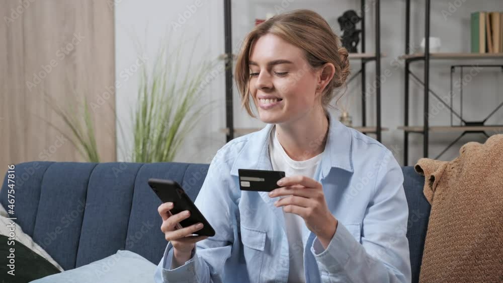 Online Shopping girl using smart phone indoor. Smiling young customer holding credit card and mobile phone sitting on couch at home. Female shopper purchase in online store. E-banking app service. Online Shopping girl using smart phone indoor. Smiling young customer holding credit card and mobile phone sitting on couch at home. Female shopper purchase in online store. E-banking app service.