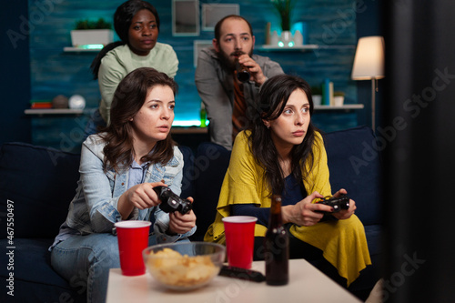 Concentrated women relaxing on couch playing video games using controller during online competition enjoying spending time together in living room. Group of multi-ethnic friends having fun at home