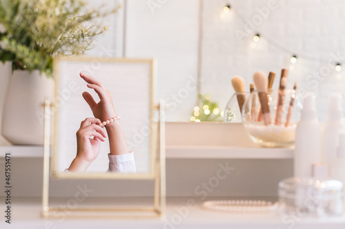 Reflection in table mirror of woman hand  in pearl bracelets. Female party preparation.   hristmas  light bulbs  eucalyptus  fir   pine branches in a vase  on dressing table with make up accessories.