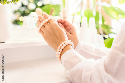 Woman puts on jewelry, pearl bracelets on the hand and  sitting near dressing table with make up accessories. Female party preparation. Reflection of christmas tree, light bulbs in table mirror.