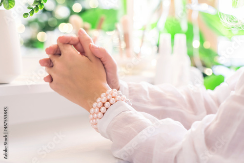 Woman hand in pearl bracelets sitting near dressing table with make up accessories. Reflection of christmas tree, light bulbs in table mirror.