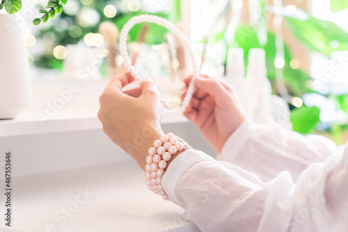Woman puts on hair band, jewelry, pearl bracelets on the hand and sitting near dressing table with make up accessories. Reflection of christmas tree, light bulbs in table mirror.