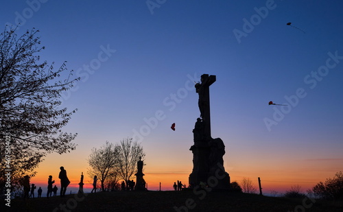 Silhouettes of kids flying kites with stone christianity monument in the foreground at sunset.