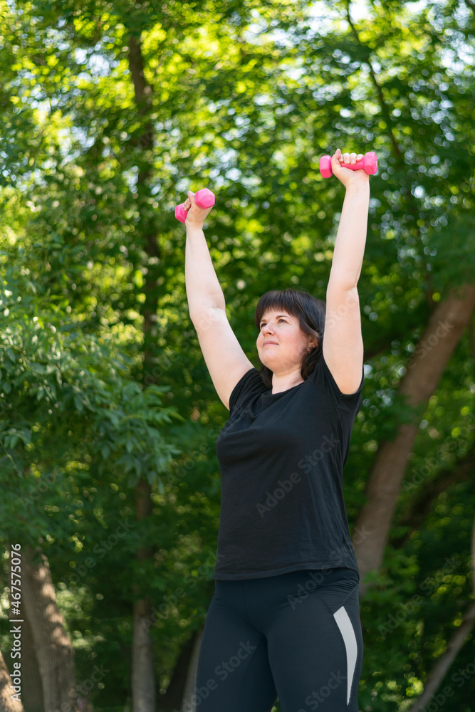 Girl coach performs exercises with dumbbells in the park. Outdoor fitness. Hand training with dumbbells. For beginners