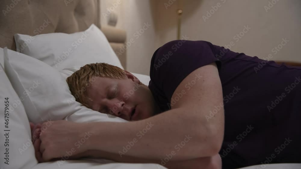 Close-up side view of exhausted adult redhead caucasian man going to sleep and lying on side on bed with white linens. Person wears purple t-shirt. Healthy night rest theme. Close-up side view of exhausted adult redhead caucasian man going to sleep and lying on side on bed with white linens. Person wears purple t-shirt. Healthy night rest theme.