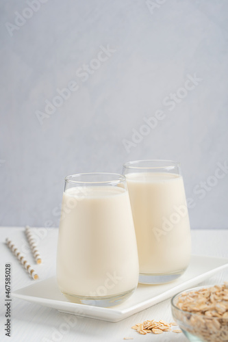 Vegetarian oat milk with creamy oatmeal taste, lactose and dairy free served in drinking glass on white wooden table for breakfast on plate with oats grain in bowl and paper straw. With copy space