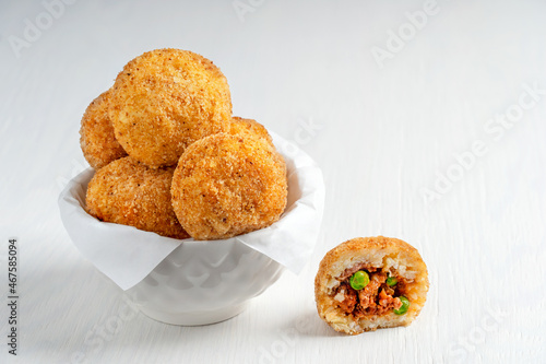 Tasty homemade Arancini Italian or Sicilian rice balls stuffed with minced meat, tomato sauce and green peas, coated with bread crumbs and deep fried, served in bowl with paper on white wooden table