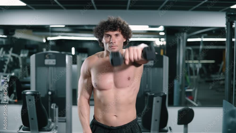 Young athletic man lifts weights in gym. Strongman raises dumbbells in front of him with outstretched arms. Front view. Young athletic man lifts weights in gym. Strongman raises dumbbells in front of him with outstretched arms. Front view.