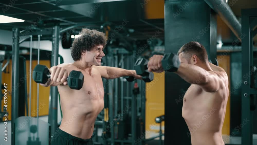 Two young athletic men doing dumbbell swings while standing opposite each other in gym. Steadicam shot. Two young athletic men doing dumbbell swings while standing opposite each other in gym. Steadicam shot.