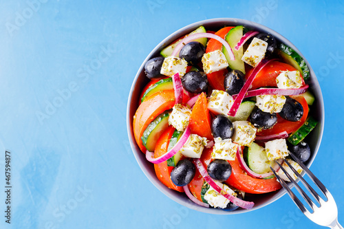 Greek salad and fork on a blue background. Fresh vegetables, feta cheese and black olives. Trendy food. Copy space. Top view
