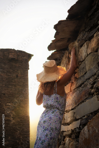 A young girl in a dress and hat stands next to the medieval towers in Svaneti at sunset. photo